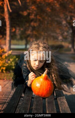Halloween. Une jolie fille aux cheveux longs se trouve sur le banc dans le parc et dessine un visage drôle à la citrouille. Banque D'Images