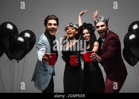 des amis multiethniques excités en costumes d'halloween tenant des tasses en plastique et des mains en agitant isolées sur le gris Banque D'Images