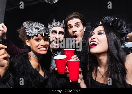 des amis multiethniques enthousiastes regardant l'appareil photo tout en dorant avec des tasses en plastique sur le noir Banque D'Images