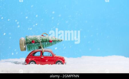 Voiture rouge portant l'arbre de noël sur le toit dans la neige sur fond bleu. Arrière-plan de Noël. Carte de vacances. Copier l'espace. Banque D'Images