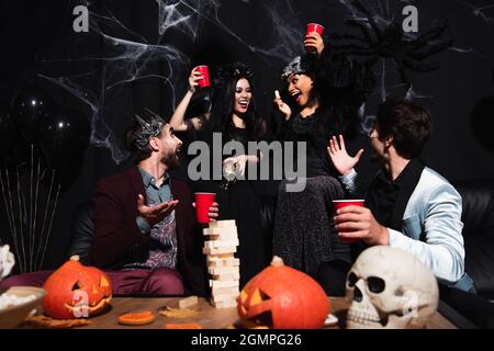 hommes pointant avec les mains près de blocs de bois jeu tandis que les femmes gaies interraciales dans les costumes d'halloween toasting avec des tasses en plastique sur noir Banque D'Images