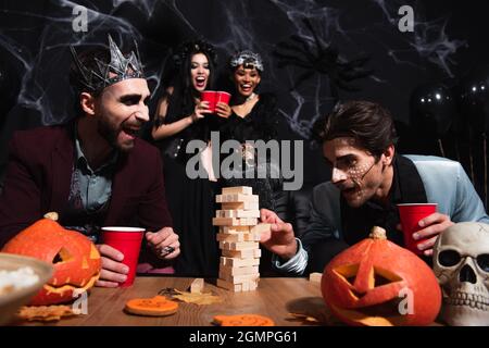 homme en halloween maquillage jouer à des blocs de bois jeu tandis que les femmes interraciales flou toaster avec des tasses en plastique sur noir Banque D'Images