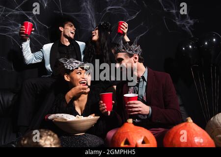 amis multiethniques en costumes d'halloween parlant tout en mangeant du pop-corn et en buvant de la bière sur le noir Banque D'Images