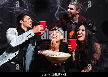 des hommes heureux en train de griller avec une tasse en plastique près des femmes interraciales en costumes d'halloween mangeant du pop-corn sur le noir Banque D'Images