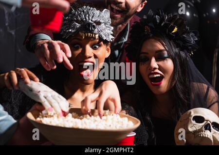 femmes interraciales excitées en costumes d'halloween, avec crâne et main de jouet près du bol avec pop-corn Banque D'Images