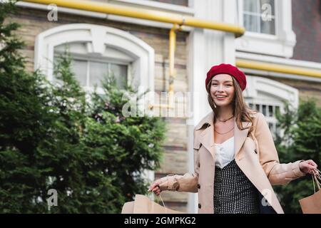 jeune femme heureuse en béret rouge et trench-coat beige tenant des sacs de shopping à l'extérieur Banque D'Images