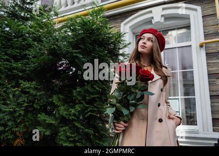 jeune femme en trench beige et béret tenant des roses rouges et debout avec la main dans la poche près du bâtiment Banque D'Images