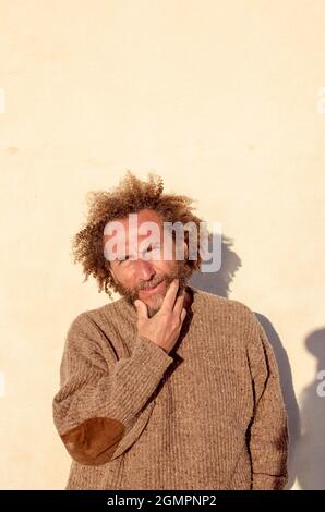 un homme d'âge moyen aux cheveux bouclés touchant sa barbe regarde l'appareil photo devant un mur blanc Banque D'Images