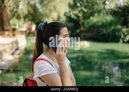 Photo moyenne d'une jeune femme avec une robe blanche et un sac à dos rouge parlant au téléphone dans un parc Banque D'Images