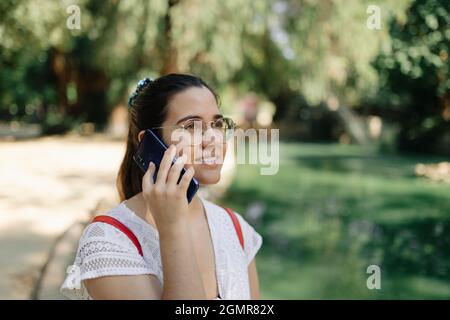 Photo moyenne d'une jeune femme avec une robe blanche et un sac à dos rouge parlant au téléphone dans un parc Banque D'Images