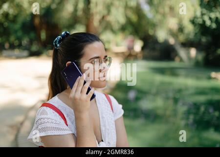 Photo moyenne d'une jeune femme avec une robe blanche et un sac à dos rouge parlant au téléphone dans un parc Banque D'Images