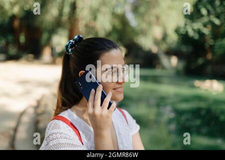 Photo moyenne d'une jeune femme avec une robe blanche et un sac à dos rouge parlant au téléphone dans un parc Banque D'Images