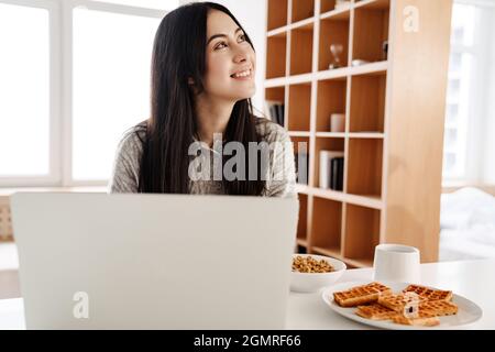 Jeune femme attrayante utilisant un ordinateur portable tout en s'asseyant dans la cuisine pour prendre le petit déjeuner Banque D'Images