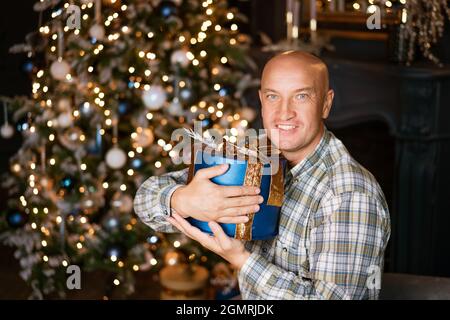 Joyeux chauve dans une chemise tenant une boîte bleue avec un cadeau sur le fond d'un arbre de noël Banque D'Images