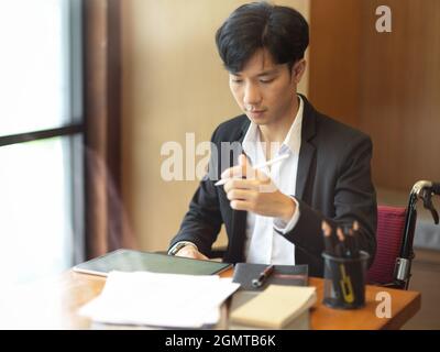 Jeune homme d'affaires en fauteuil roulant travaillant à son bureau, travaillant sur une tablette numérique, fournitures de bureau sur une table Banque D'Images