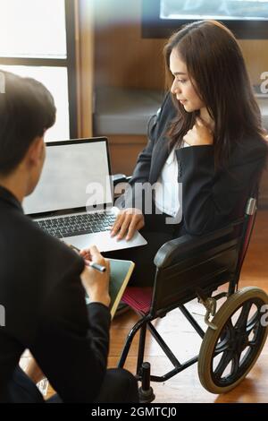 Jeune femme d'affaires en fauteuil roulant avec ordinateur portable mockup consultant avec un collègue au bureau Banque D'Images