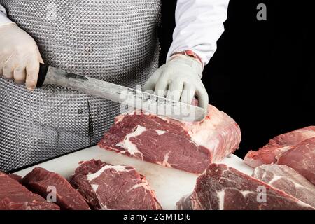 Gros plan des morceaux de viande hachés et des mains de boucher dans des gants spéciaux coupant avec un couteau. Viande de porc ou de bœuf sur la table du boucher. Le travailleur en tout Banque D'Images