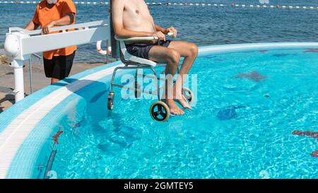 Homme handicapé à la piscine ascenseur pour fauteuil roulant et transféré dans l'eau par un assistant. Banque D'Images