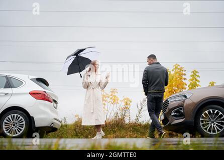 Un jeune couple sur la route à proximité de voitures dans un ciel nuageux avec pluie, se disant Au revoir l'un à l'autre. Banque D'Images