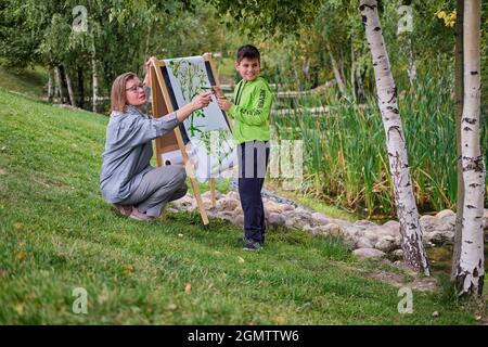 Mère enseigne garçon fils à peindre sur toile. Femme enseignante artiste peint avec un enfant sur la nature papier et les arbres au bord de la rivière Banque D'Images