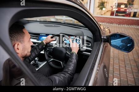 Beau jeune homme qui choisit une nouvelle voiture à la concession, assis à l'intérieur d'une voiture. Banque D'Images