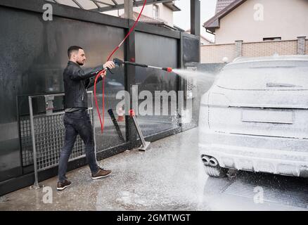 Homme élégant debout au poste de lavage de voiture, tenant un pistolet spécial pour appliquer la mousse à auto sous haute pression. Le lavage de l'automobile avec un détergent sous pression est la première étape du nettoyage professionnel de la voiture. Banque D'Images