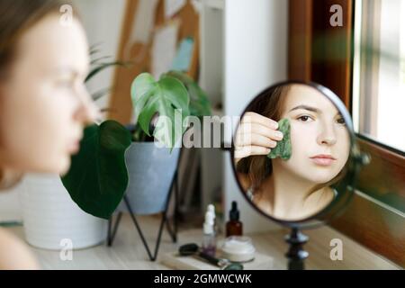 Gros plan portrait de la femme caucasienne adulte, utilisant le rouleau de pierre de gua sha jade naturel pour masser le visage dans la journée à la maison. Espace de photocopie avec vue avant des cosmétiques et soins de beauté. Banque D'Images