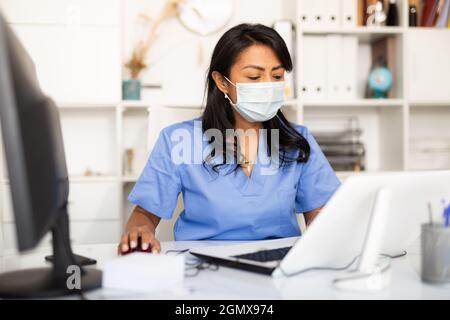 Femme expérimentée médecin dans le masque de protection remplissant les formes médicales sur ordinateur portable tout en étant assis à la table dans le bureau Banque D'Images
