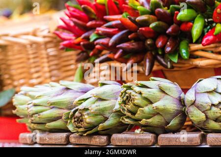 Artichauts verts frais avec des feuilles vertes et des petits pains de piments rouges et verts chauds dans un panier dans un marché alimentaire de rue Banque D'Images