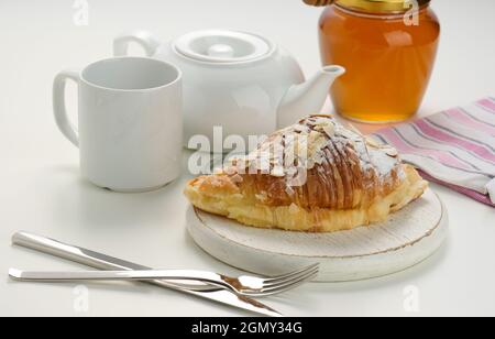 Un croissant croustillant cuit est arrosé de sucre en poudre et de flocons d'amandes sur un panneau de bois, une infusion de céramique blanche et une tasse sur une table blanche. Petit déjeuner Banque D'Images