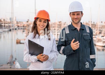 Jeunes hommes et femmes joyeux gestionnaires logistiques en casques et uniforme de protection avec presse-papiers et radio VHF regardant la caméra tout en contrôlant l'expédition dans le port de mer Banque D'Images
