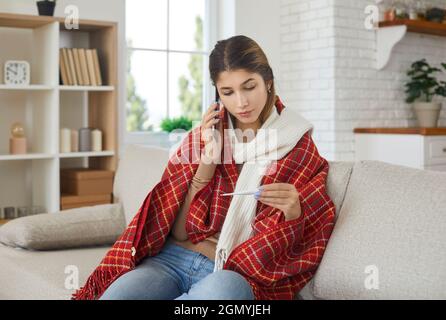 Une jeune femme malade parle au téléphone portable et vérifie le thermomètre de maintien de température. Banque D'Images