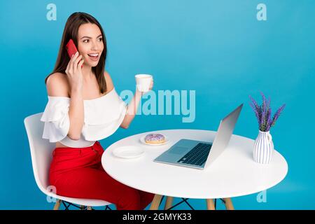 Portrait d'une jolie fille gaie assise à table mangeant des bonbons buvant un espresso parlant au téléphone isolé sur fond bleu clair Banque D'Images