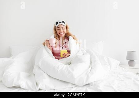 Drôle millénaire blanche blonde femme en pyjama rose et masque de sommeil aime le café et lit le message sur smartphone Banque D'Images