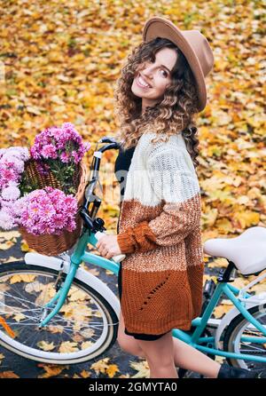 Vue latérale d'une brunette en forme de curly voyageant en vélo vintage avec un grand panier de fleurs roses. Jolie femme portant un chapeau marron et un gilet est à vélo dans le parc d'automne. Concept de week-end . Banque D'Images