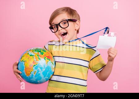 Photo de jeune petit garçon blond tenir main ID tag globe leçon sourire isolé sur fond rose couleur Banque D'Images