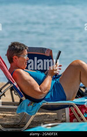 une femme d'âge moyen âgée lit sur un kindle ou un appareil de lecture électronique alors qu'elle était sur un transat sur la plage en vacances en grèce. Banque D'Images