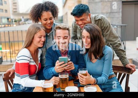 Un groupe de jeunes amis variés se sont rassemblés autour d'un smartphone à une table de pub pour apprécier une bière en riant et en plaisantant pendant qu'ils font un appel vidéo Banque D'Images