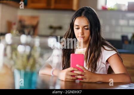 Une jeune adolescente attrayante lit ou regarde des médias sur son téléphone mobile pendant qu'elle est assise à l'intérieur se détendant à une table Banque D'Images