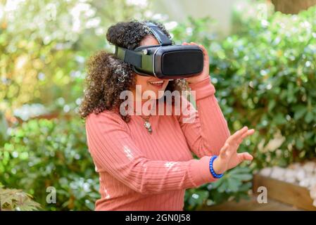 Jeune femme utilisant un micro-casque de réalité virtuelle 3D en plein air sur un patio en faisant des gestes avec sa main alors qu'elle s'immerge dans son environnement simulé contre Banque D'Images