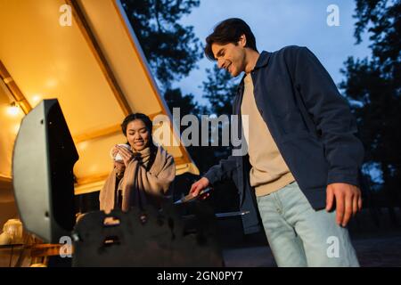 Vue en contre-plongée d'un homme souriant qui cuisine sur le grill près d'une petite amie asiatique avec une tasse et une maison de luxe à l'extérieur Banque D'Images