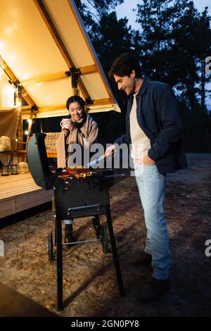 Couple interracial souriant avec une tasse de cuisson au grill sur une terrasse de luxe Banque D'Images