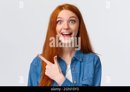 Gros plan portrait excité Happy Ginger fille, cheveux foxy et yeux bleus, sourire optimiste avec étonnement et joie, pointage doigt gauche, assister à l'amusant cool Banque D'Images