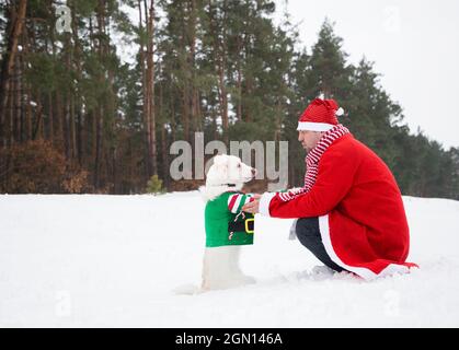 Un homme en costume rouge du Père Noël et un chien blanc en veste verte sont assis l'un en face de l'autre sur le sol enneigé. Bonjour hiver, joyeux noël Banque D'Images