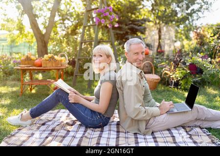 Heureux couple senior reposant sur une couverture de pique-nique dans le jardin, homme utilisant un ordinateur portable, femme lisant le livre Banque D'Images