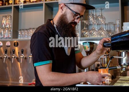 Barista barbu qui prépare de la crème de lait cuite à la vapeur dans un pot en acier, à l'aide d'une machine à café professionnelle. Préparation d'un café Latte ou d'un cappuccino. Banque D'Images