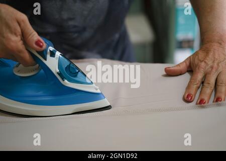 Une femme âgée se tient à la main tout en repassant des vêtements avec un fer à vapeur sur une planche à repasser Banque D'Images