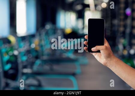 Homme main tenant smartphone avec écran noir vierge sur fond intérieur de salle de gym Banque D'Images