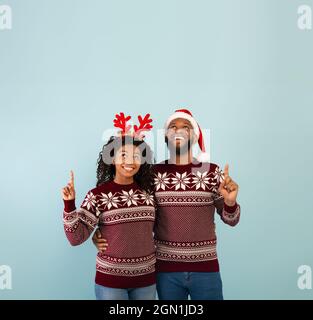 Offre vacances. Heureux conjoints noirs dans des chandails identiques de Noël pointant vers le haut de l'espace de copie, fond bleu Banque D'Images