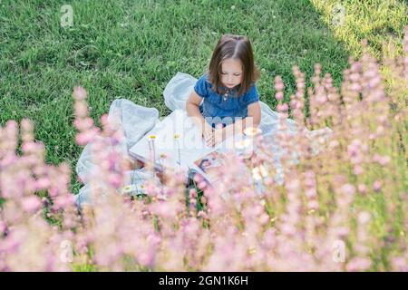 Petite fille partiellement floue 3-4 avec des cheveux foncés dans la robe de denim s'assoit et lit livre sur la pelouse près des fleurs Banque D'Images